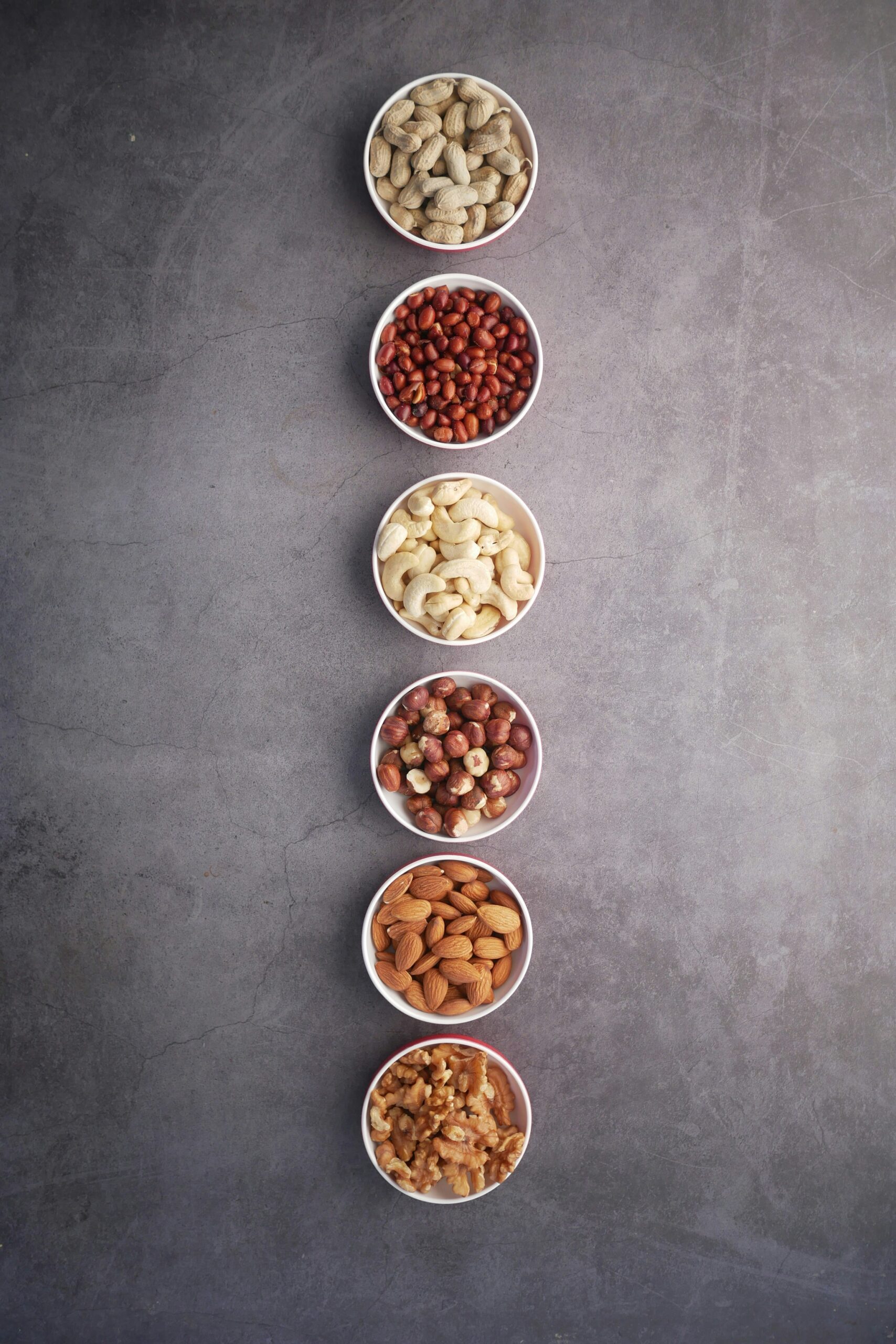 Top down view of six small bowls, sitting on a grey stone surface, each filled with a different type of nuts